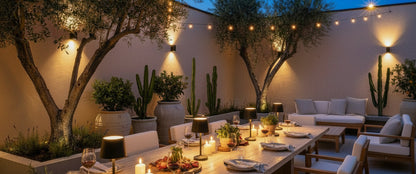 An outdoor dining patio at dusk with a long wooden table set for six, candles, string lights, olive trees, cacti, and a lounge sofa in the background.